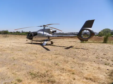 Helicopter standing on dry ground beside vineyard rows under a clear sky