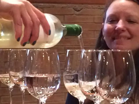 Hand pouring white wine into a line of tasting glasses, host smiling behind
