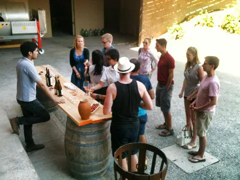 Group of guests tasting wine at a plank table set on barrels in a winery courtyard, with a host pouring