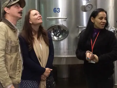 Winery guide with a lanyard talking to two guests in front of a numbered stainless steel fermentation tank